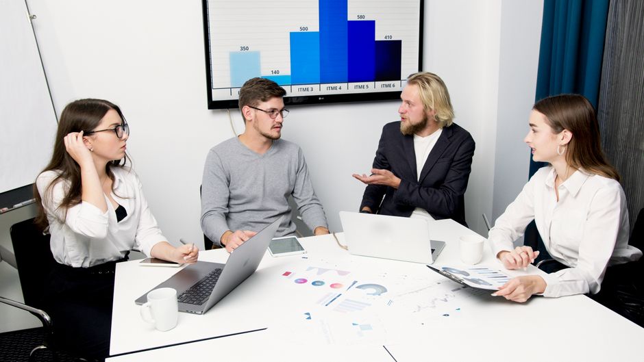 A group of business professionals discussing data at a meeting table with laptops and charts.